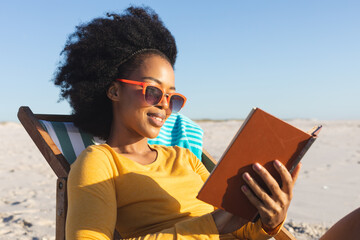 Happy african american woman in sunglasses sitting in deckchair reading book on sunny beach