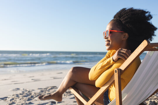 Happy African American Woman In Sunglasses Sitting In Deckchair Smiling On Sunny Beach By Sea