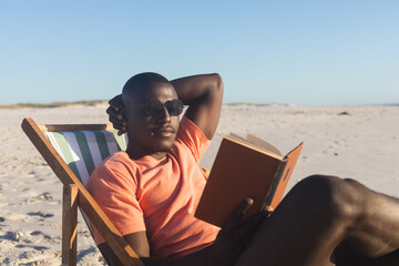 Happy african american man in sunglasses sitting in deckchair reading book on sunny beach