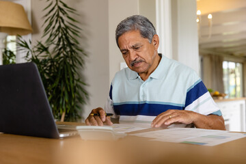 Senior biracial man doing paperwork and using laptop in dining room