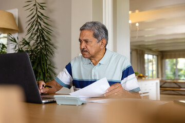 Senior biracial man doing paperwork and using laptop in dining room