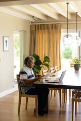 Biracial senior man doing paperwork and using laptop in dining room