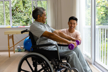 Happy diverse male physiotherapist advising and senior male patient in wheelchair using dumbbells