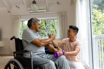 Happy diverse male physiotherapist advising and senior male patient in wheelchair using dumbbells