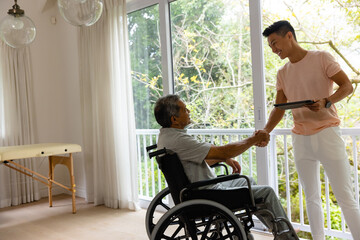 Happy diverse male physiotherapist shaking hands with senior male patient in wheelchair
