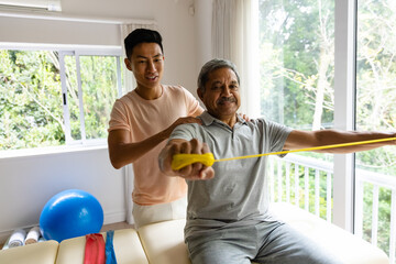 Happy diverse male physiotherapist advising and senior male patient using exercise band