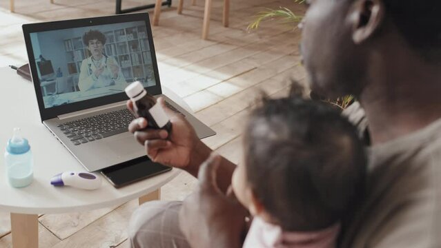 Over the shoulder shot of black dad sitting with sick baby at home, holding syrup and speaking with female doctor during online consultation via video call on laptop