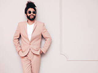 Handsome smiling hipster  model. Unshaven Arabian man dressed in elegant beige suit . Fashion male with long curly hairstyle posing near grey wall in studio. Isolated. In sunglasses