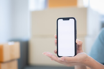 Delivery app. Close-up of smiling young woman in hand showing smartphone with blank screen for mockup woman working with boxes at home