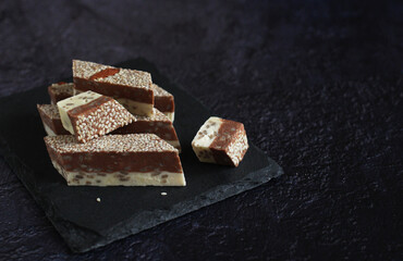 milk-chocolate halva with sesame seeds and nuts in pieces on a board on a dark background