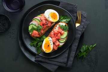 Healthy sandwich with bread or toast, smoked salmon, soft egg, cream cheese, cucumber, radish, black sesame and parsley on black plate. Delicious protein fish sandwich for breakfast. Smorrebrod.