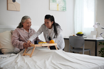 Caregiver putting tray with breakfast on bed of senior woman