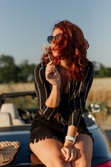 Beautiful young woman in sunglasses sits on a black convertible in a field