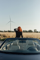 A  beautiful young woman sits on a black convertible in a field near windmills