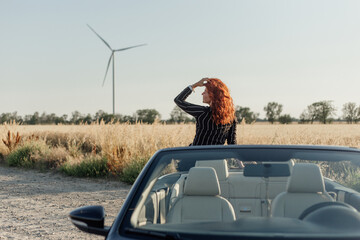 A beautiful young woman sits on a black convertible in a field near windmills