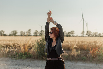 A beautiful young woman in a black leather jacket stands in a field near windmills