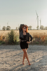 A beautiful young woman in a black leather jacket stands in a field near windmills