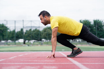 Fototapeta premium Concentrated athlete standing in low start pose before sprint on red rubberized track surface on city sports ground sportsman exercising at outdoor arena