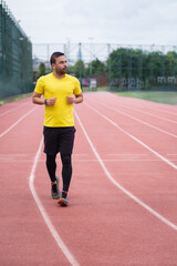 Fototapeta premium Man in tracksuit running along track with red rubber covering and white lines on outdoor sports arena in town park for health and body muscles development