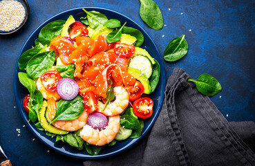 Fresh salad for keto diet with salmon, avocado, spinach, cucumber, shrimps, tomato, cashew, sesame seeds. Low-carbohydrate lunch rich in healthy fats. Blue stone table background, top view
