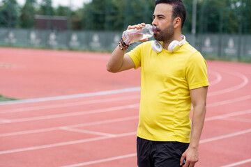 Thirsty sportsman with wireless headphones drinking water from plastic bottle and refreshing at training on running track at sports arena man keeping body balance - obrazy, fototapety, plakaty