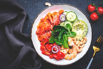 Keto diet salad with salmon, shrimp, avocado, spinach, cucumber, tomato, cashew, sesame. Low-carbohydrate lunch rich in healthy fats. Black table background, top view