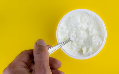 Top view of a person's hand holding a spoon in a yogurt against a yellow background.
