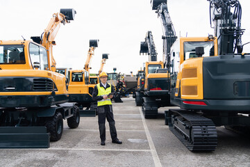 Engineer in a helmet with a digital tablet stands next to construction excavators.