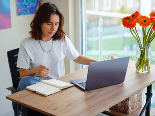 Beautiful Latin young woman in casual clothing using laptop and smiling while working indoors. lady work on computer listening music with earphones.