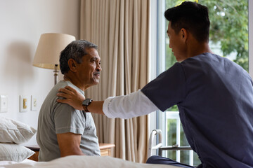 Diverse male doctor and senior male patient sitting on bed and discussing at home