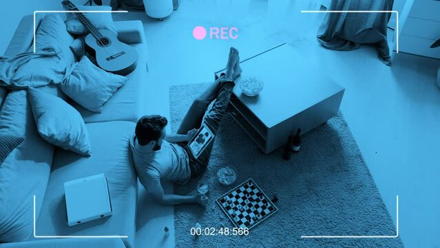 High angle spy camera shot of young man sitting on floor playing chess and drinking beer with his friend during video call on digital tablet