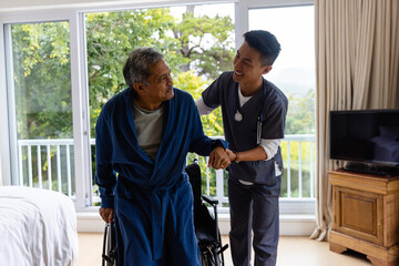 Diverse male doctor helping senior male patient getting up from wheelchair at home