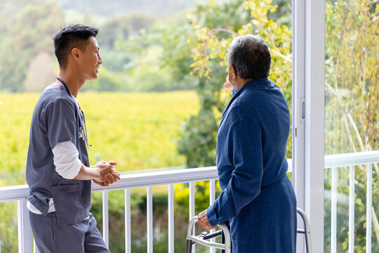 Diverse Male Doctor And Senior Male Patient Using Crutches Looking Out Window At Home