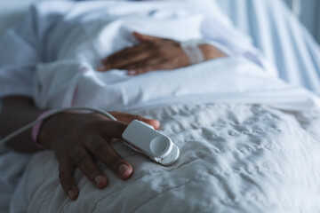 Obraz na płótnie Canvas Midsection of african american female patient with pulse oximeter on hand, lying on bed at hospital