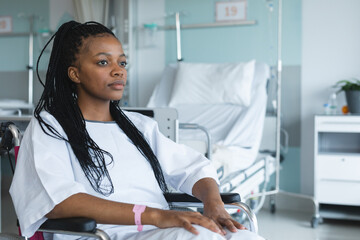 African american female patient wearing hospital gown, sitting in wheelchair in hospital room