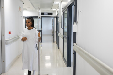 African american female patient wearing hospital gown, holding drip and walking in corridor