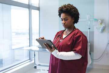 African american female doctor wearing scrubs and stethoscope, using tablet in hospital room