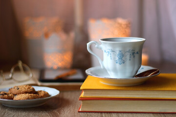 Cup of tea or coffee, plate of cookies, books, e-reader, pencil and lit candles on the table. Selective focus.