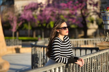 Fototapeta premium Young and beautiful woman, Hispanic, brown hair, wearing striped sweater, linen pants and sunglasses, leaning on a railing, looking straight ahead relaxed and calm. Concept relax, peace, tranquility.