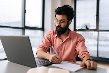 Portrait of bearded Indian businessman in glasses using laptop analyzing working project results, writing notes siting at table by window. Focused student male learning remotely looking to screen.