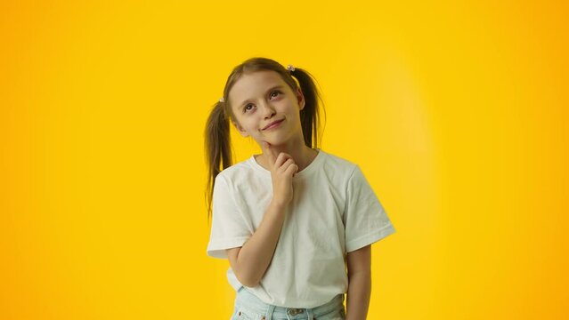 Young girl thinking and dreamy smiling while touching her chin with a finger on a yellow background. Concept of childhood dreams of a successful future