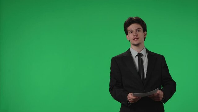 Reporting TV presenter with papers in his hands in a studio against a green screen. Professional Anchorman speaks confidently and points to the green screen. Advertising area, workspace mock up.