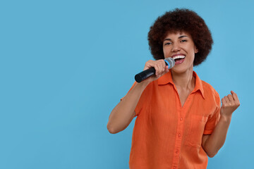 Curly young woman with microphone singing on light blue background, space for text