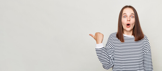 Surprised astonished brown haired adult woman wearing striped shirt standing isolated over gray background looking at camera with open mouth pointing at copy space.