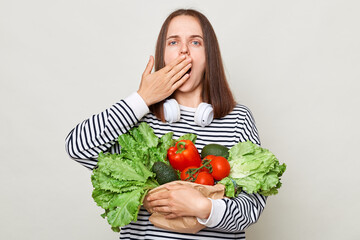 Shocked surprised brown haired young woman embraces bouquet of fresh vegetables wearing striped casual shirt isolated over gray background covering mouth with palm.