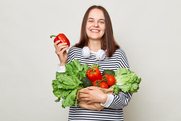 Satisfied delighted brown haired young woman embraces bouquet of fresh vegetables wearing striped casual shirt isolated over gray background showing red pepper.