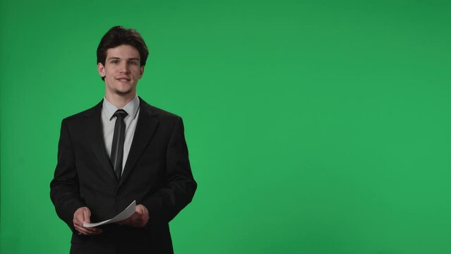 Reporting TV presenter in the studio against the background of a green screen. A male TV presenter with papers in his hands confidently speaks, listens, nods. Advertising area, workspace mock up.