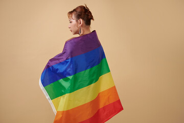 Serious transgender woman wrapped in rainbow flag standing against beige background