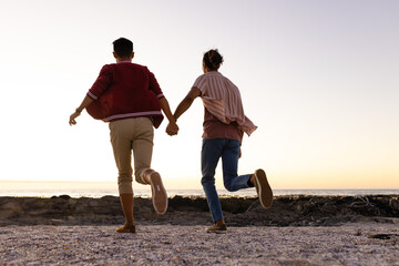Happy biracial gay male couple holding hands and running on beach at sunset