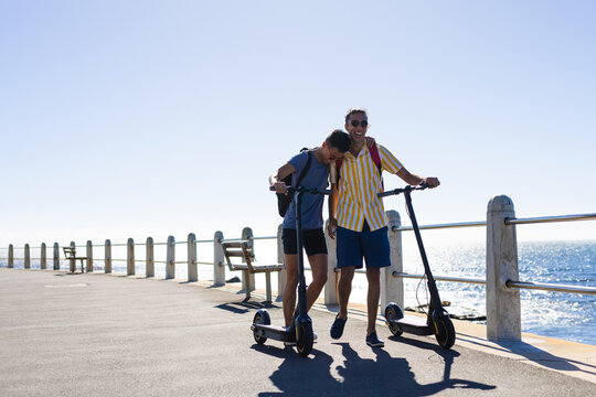 Happy biracial gay male couple walking with scooters on promenade by the sea, copy space